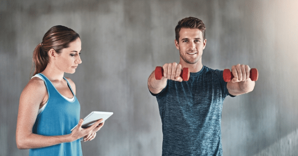 A woman with a tablet stands on the left, observing a young man in his 20s lifting red dumbbells in front of a gray wall.