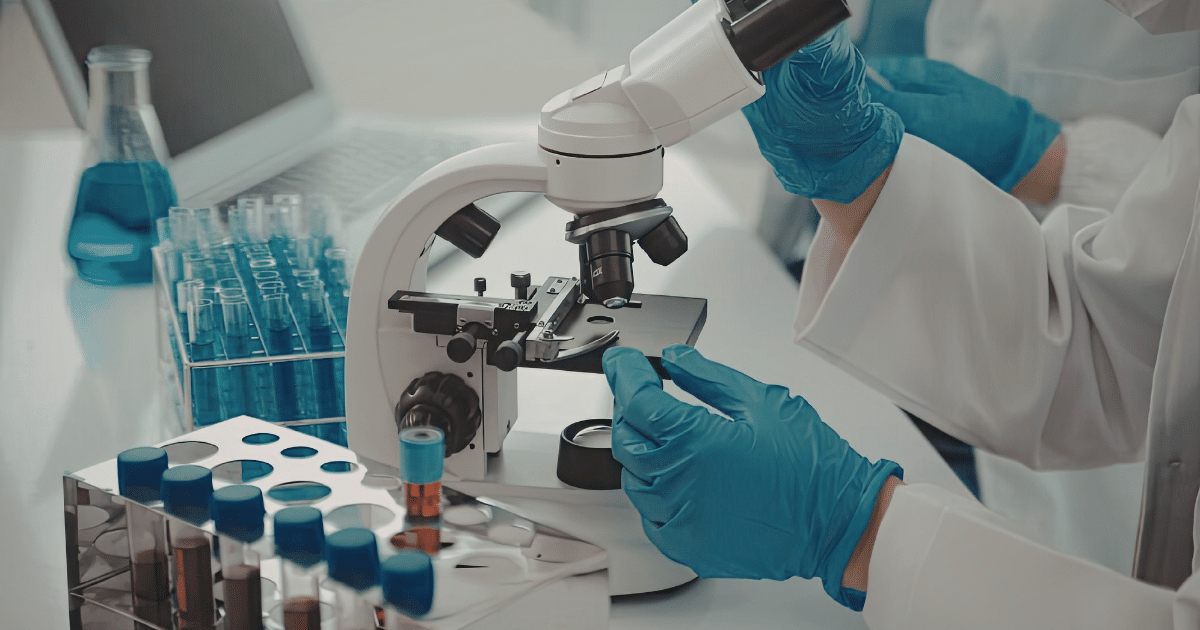 A close-up view of a scientist's gloved hands operating a white laboratory microscope, with test tube racks and beaker equipment set on the desk nearby.