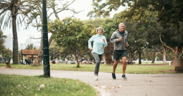 An active senior couple, a woman in a light blue jacket and a man in a grey windbreaker, jogging together along a paved path in a lush green park on an overcast day.