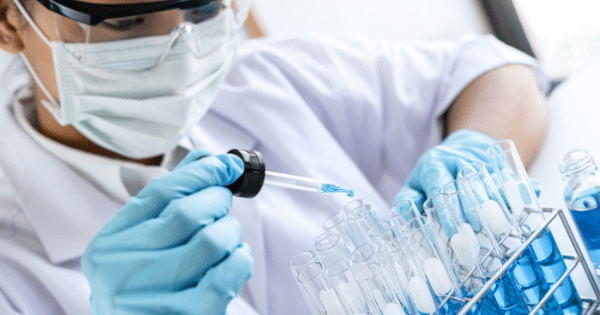 Close-up of a scientist wearing a white lab coat, medical mask, protective goggles, and blue gloves. They are using a glass dropper to carefully dispense a blue liquid into a row of test tubes arranged in a metal rack.