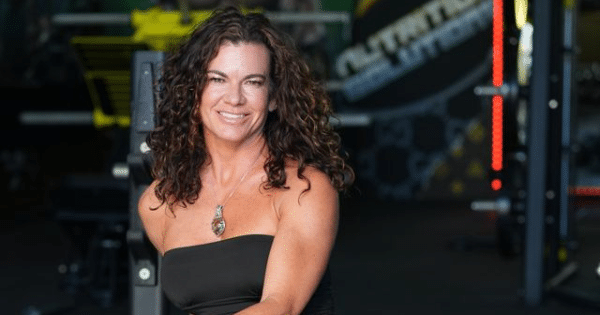 A middle-aged woman with long, curly brown hair and a bright smile, wearing a black strapless top and a pendant necklace. She is posing in a softly lit gym environment with fitness equipment blurred in the background.