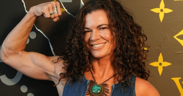 A smiling woman with dark, curly hair wearing a denim top and a large green pendant necklace, flexing her right bicep to showcase muscle definition.