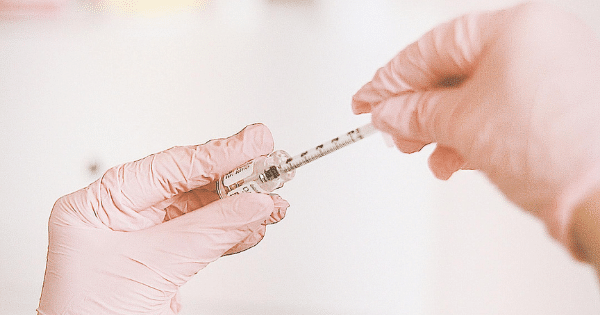 Hands wearing pink nitrile gloves holding a small liquid medication vial and drawing a dose into a fine-gauge syringe.