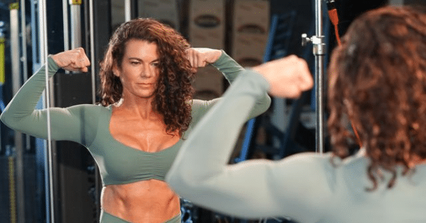 A fit woman with long curly hair wearing a green athletic crop top, flexing both arms in front of a mirror at a gym while looking at her reflection.