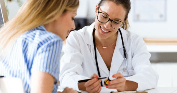 A smiling female doctor wearing glasses, a white coat, and a stethoscope pointing a pen at a small prescription medication bottle while speaking with a patient.