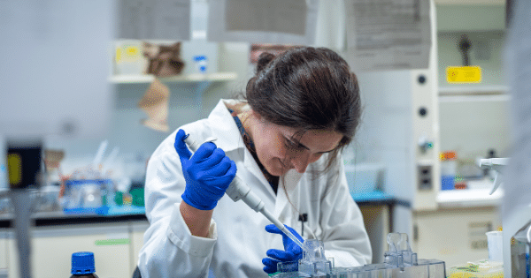 A female scientist wearing a white lab coat and blue protective gloves carefully using a precision micropipette to transfer liquid into a lab container in a research laboratory setting.