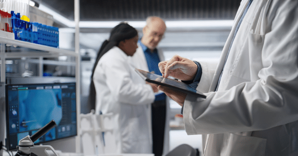 A close-up view of a scientist in a white lab coat operating a digital tablet in a high-tech laboratory, while two colleagues discuss research in the blurred background.