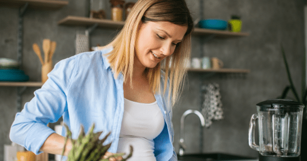 A blonde woman in a blue open button-down shirt preparing fresh asparagus on a kitchen counter next to a blender.