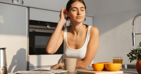 A woman sitting at a sunlit kitchen counter with her eyes closed, enjoying the warm sunlight, beside a cup of coffee, sliced citrus fruits, fresh juice, and a newspaper.