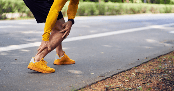A runner bent over on a paved road holding their lower calf muscle in pain due to a leg injury or cramp, wearing bright yellow athletic shoes.