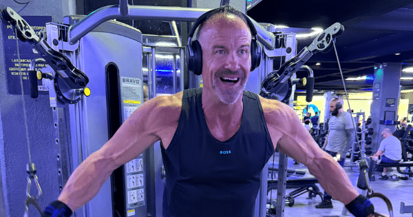 A fit, muscular middle-aged man with headphones smiling as he performs a cable chest fly exercise on a gym machine.