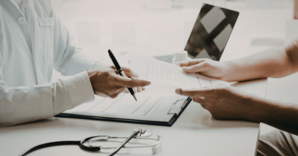 A doctor in a white coat handing medical paperwork and a prescription form across a desk to a patient, with a stethoscope sitting in the foreground.