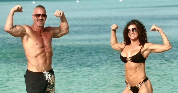 A athletic couple in swimwear standing in tropical turquoise ocean water, smiling and flexing their biceps together.
