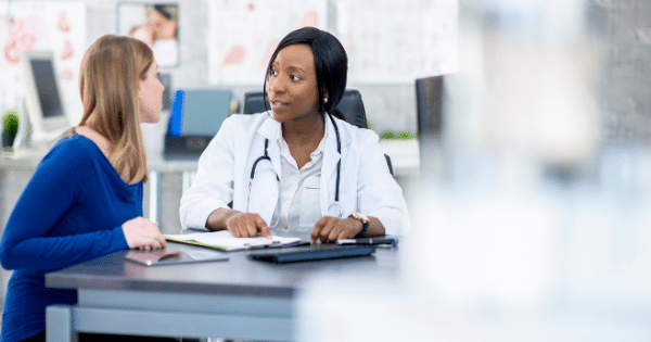 A female doctor in a white coat sits at her desk, pointing at paperwork while discussing and consulting with a female patient sitting opposite her.