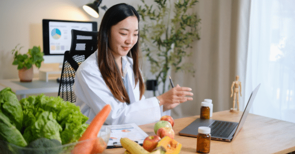 A female nutritionist in a white coat sits at a wooden desk with a laptop, pills, and fresh produce like lettuce, carrots, apples, and bananas, talking during a virtual consultation.
