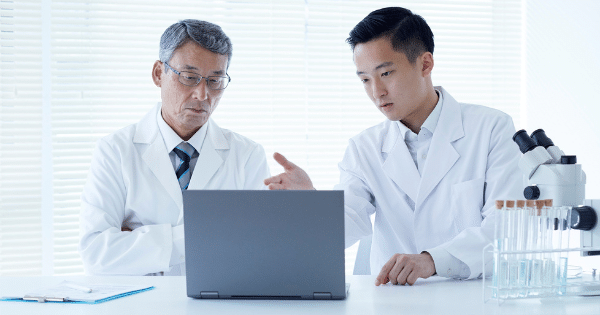 Two Asian medical researchers in white lab coats—one older with glasses, one younger—collaborating over a laptop at a laboratory desk next to a microscope and test tubes.