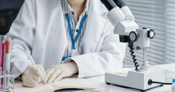 A researcher in a white lab coat and protective gloves writing notes in an open journal next to a white laboratory microscope and petri dishes.