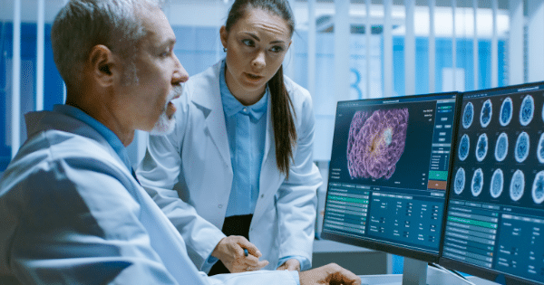 An older male doctor and a younger female medical professional in lab coats standing together, deeply analyzing high-resolution brain scans on dual computer monitors.