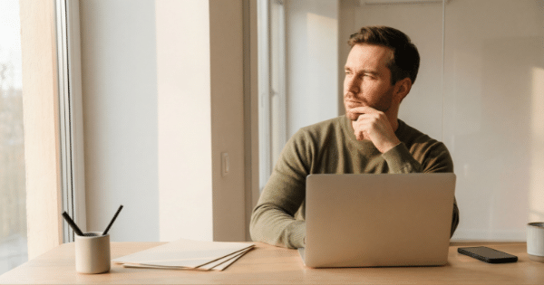 A man sitting at a wooden desk with a laptop, resting his hand on his chin while looking thoughtfully out a window.