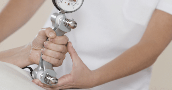 Close-up of a person gripping a silver mechanical hydraulic hand dynamometer while a practitioner rests a supporting hand underneath during a grip strength evaluation.