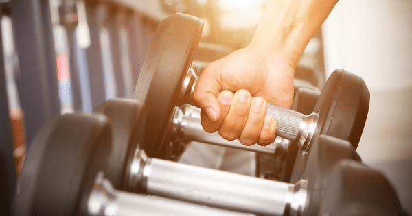 Close-up of a hand lifting a heavy chrome and black dumbbell from a weight rack in a brightly lit gym.