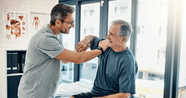 Physical therapist assisting an older man with arm mobility exercises in a clinic.