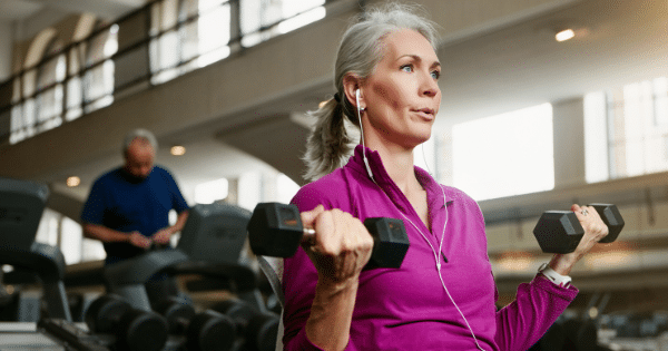 A middle-aged woman with gray hair wearing a purple long-sleeve athletic shirt and earphones performing bicep curls with dumbbells inside a fitness facility.