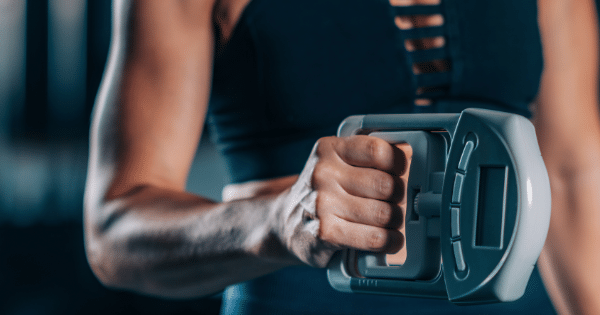 Close-up of a person's muscular forearm as they tightly squeeze a gray digital grip dynamometer in a gym setting.