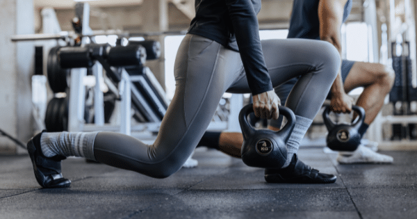 Side view of a woman and man side-by-side in a low lunge position holding black kettlebells during a workout inside a modern gym.
