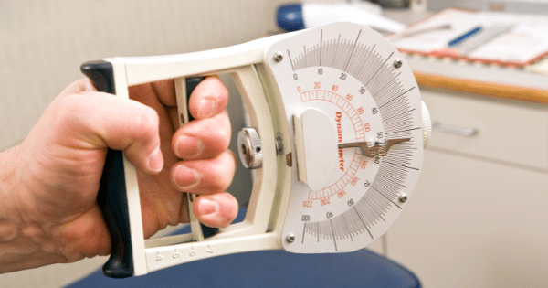 Close-up of a hand squeezing a white analog hand dynamometer with a round dial measurement gauge in a medical office setting.