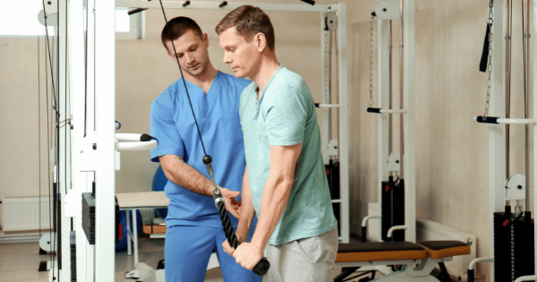A physical therapist in blue scrubs assisting a man in a light blue shirt performing a tricep cable pushdown exercise on a cable weight machine.