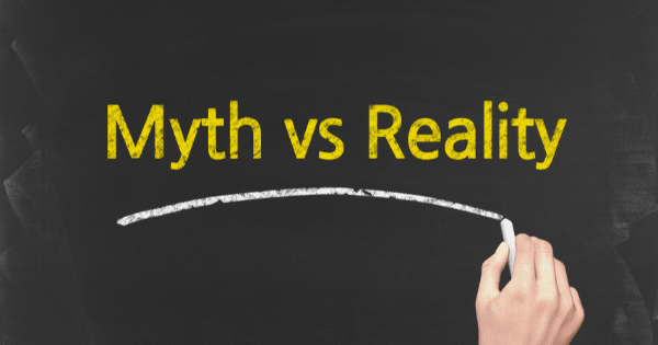 A hand holding a piece of white chalk, writing the text "Myth vs Reality" on a dark chalkboard.