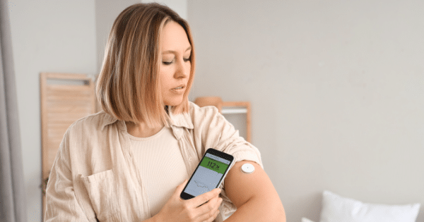 A blonde woman holding a smartphone that displays a glucose level reading of 112 next to a round white continuous glucose monitor sensor attached to her upper arm.