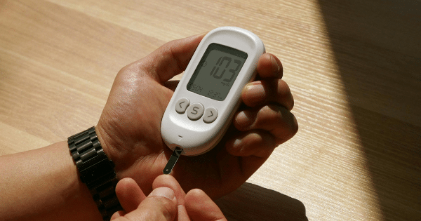Close-up of a person holding a digital blood glucose meter displaying a reading of 103 mg/dL while applying a blood drop from their fingertip to the test strip.