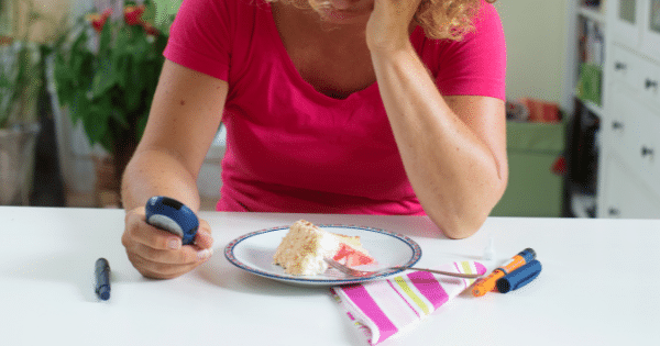 A woman sitting at a white table resting her head on her hand, looking down at a blood glucose meter next to a slice of cake, a fork, and an insulin pen.