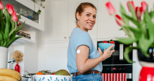 A smiling woman standing in a brightly lit kitchen holding a bowl of fresh strawberries, wearing a light blue top that reveals a white continuous glucose monitor sensor on her upper arm.