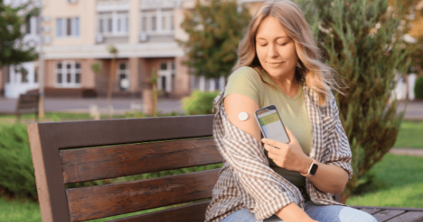 A woman sitting on an outdoor park bench, holding a smartphone showing a glucose reading next to a white continuous glucose monitor sensor on her arm.
