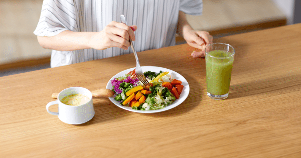 A high-angle view of a person eating a colourful mixed vegetable salad with a fork at a wooden table, alongside a mug of soup and a glass of green juice.