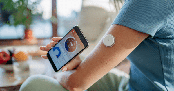Close-up of a woman holding a smartphone displaying a health monitoring app with a reading of 99, showcasing a round white continuous glucose monitor patch on her upper arm.