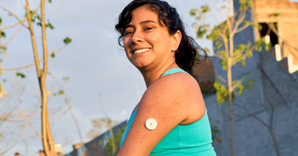 A smiling young woman wearing a bright turquoise athletic tank top outdoors, displaying a round white continuous glucose monitor sensor attached to her upper arm.