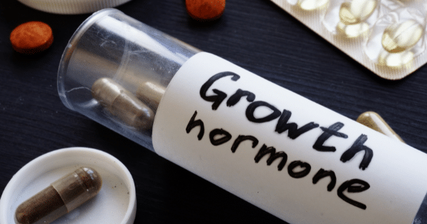 Close-up of a clear plastic bottle labeled "Growth hormone" in black handwritten text, surrounded by brown capsules, blister packs of softgels, and orange pills on a dark surface.