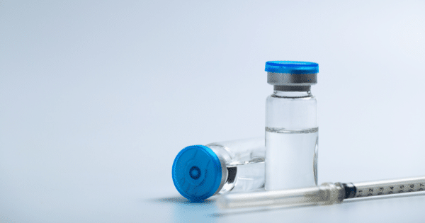 Two small medical glass vials filled with clear liquid and topped with blue caps, positioned next to a medical syringe on a pale background.