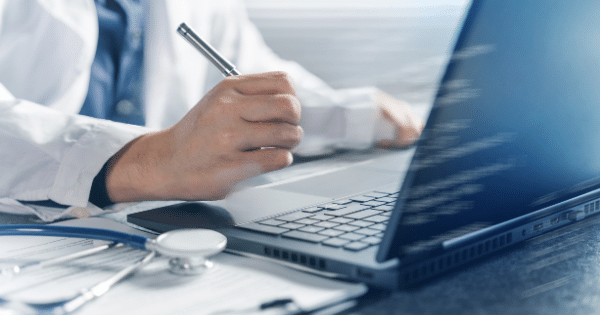Close-up of a medical professional in a white lab coat holding a pen over a desk, next to an open laptop and a stethoscope lying on medical paperwork.