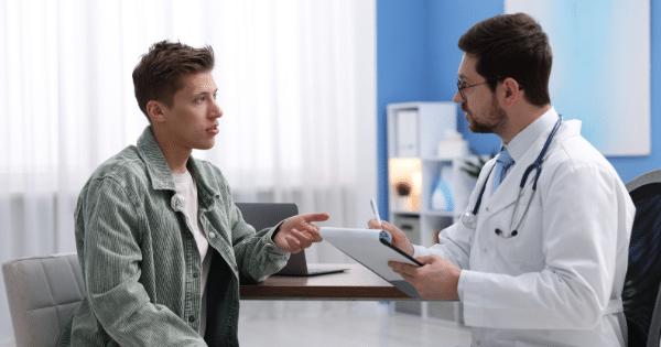 A young man in a green jacket speaking and gesturing with his hand during a medical consultation with a male doctor wearing a white coat, glasses, and a stethoscope holding a clipboard.
