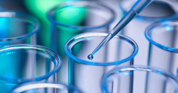 Extreme close-up of a blue-lit pipette tip dispensing a single tiny liquid drop above glass lab equipment.