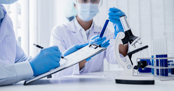 Two lab technicians wearing masks and gloves taking notes on a clipboard while analyzing a blue liquid test tube next to a microscope.