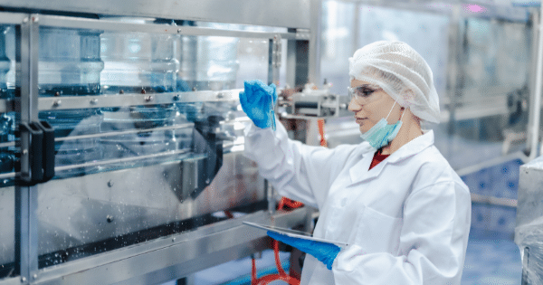 A female scientist in a hairnet, face mask, and lab coat inspecting a test tube while holding a tablet in front of industrial bottling machinery.