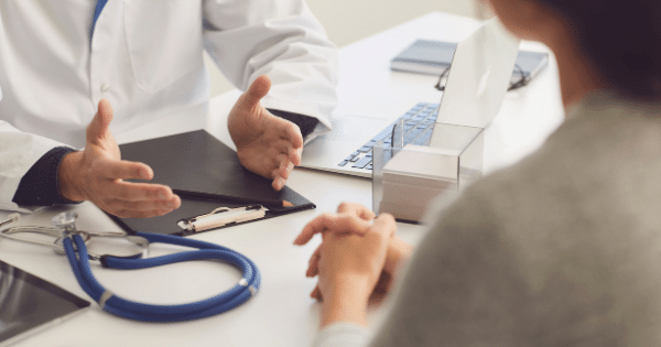 A doctor in a white coat gesturing with open hands while speaking to a patient across a desk, with a blue stethoscope and laptop nearby.