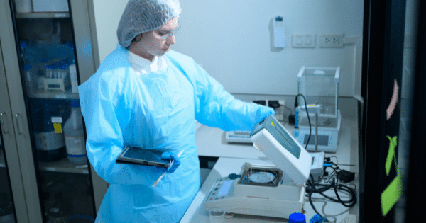 A laboratory technician in a full protective blue gown, hairnet, and safety glasses using a digital analytical scale while holding a tablet.