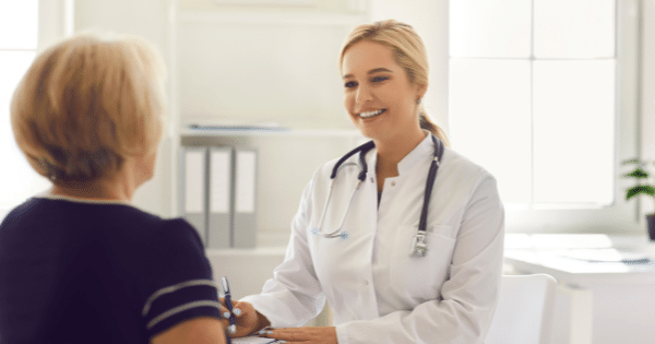 A smiling female doctor wearing a white lab coat and stethoscope sitting across from an elderly female patient during an office visit.